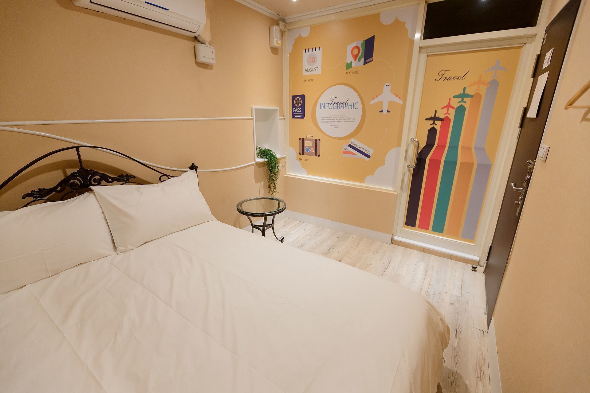 A hotel room with a bed, a small table with a plant, a wall with travel-themed decorations, and a door with a colorful design.