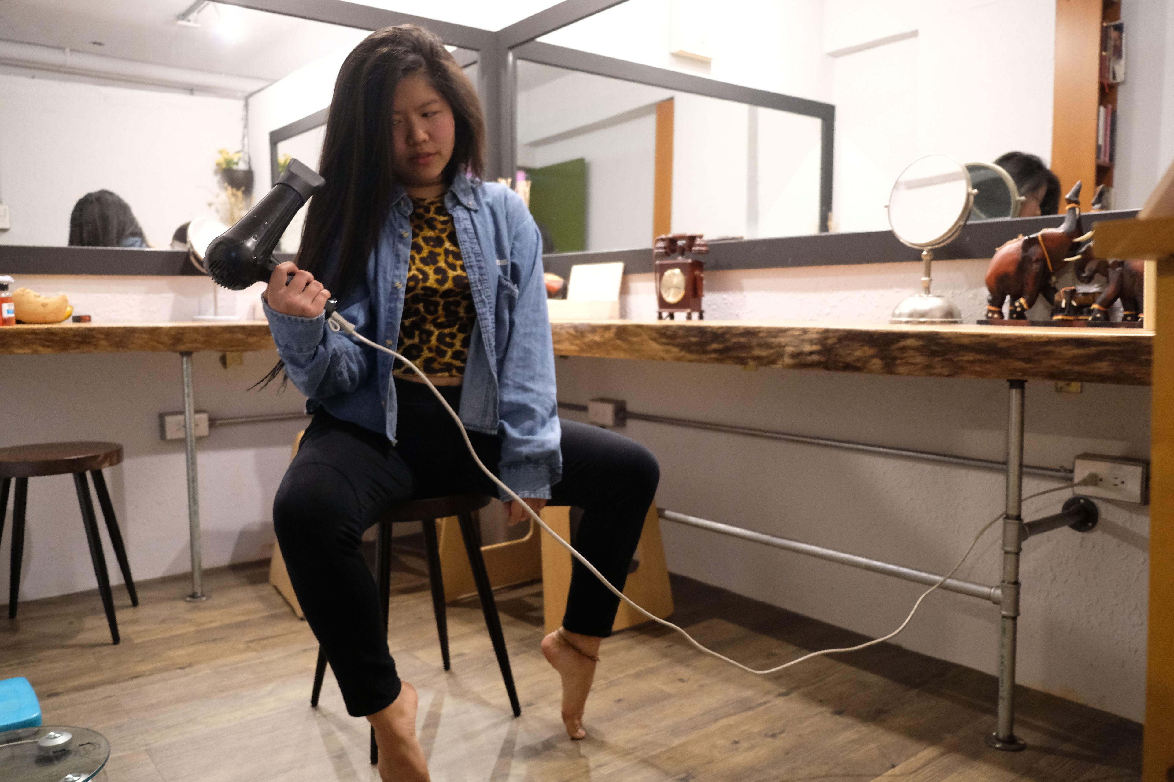 A person seated on a chair in a room with a wooden floor, a mirror, and a shelf with decorative items.