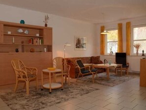 A living room with a sofa, chairs, a coffee table, a bookshelf, a painting, and a window with curtains.