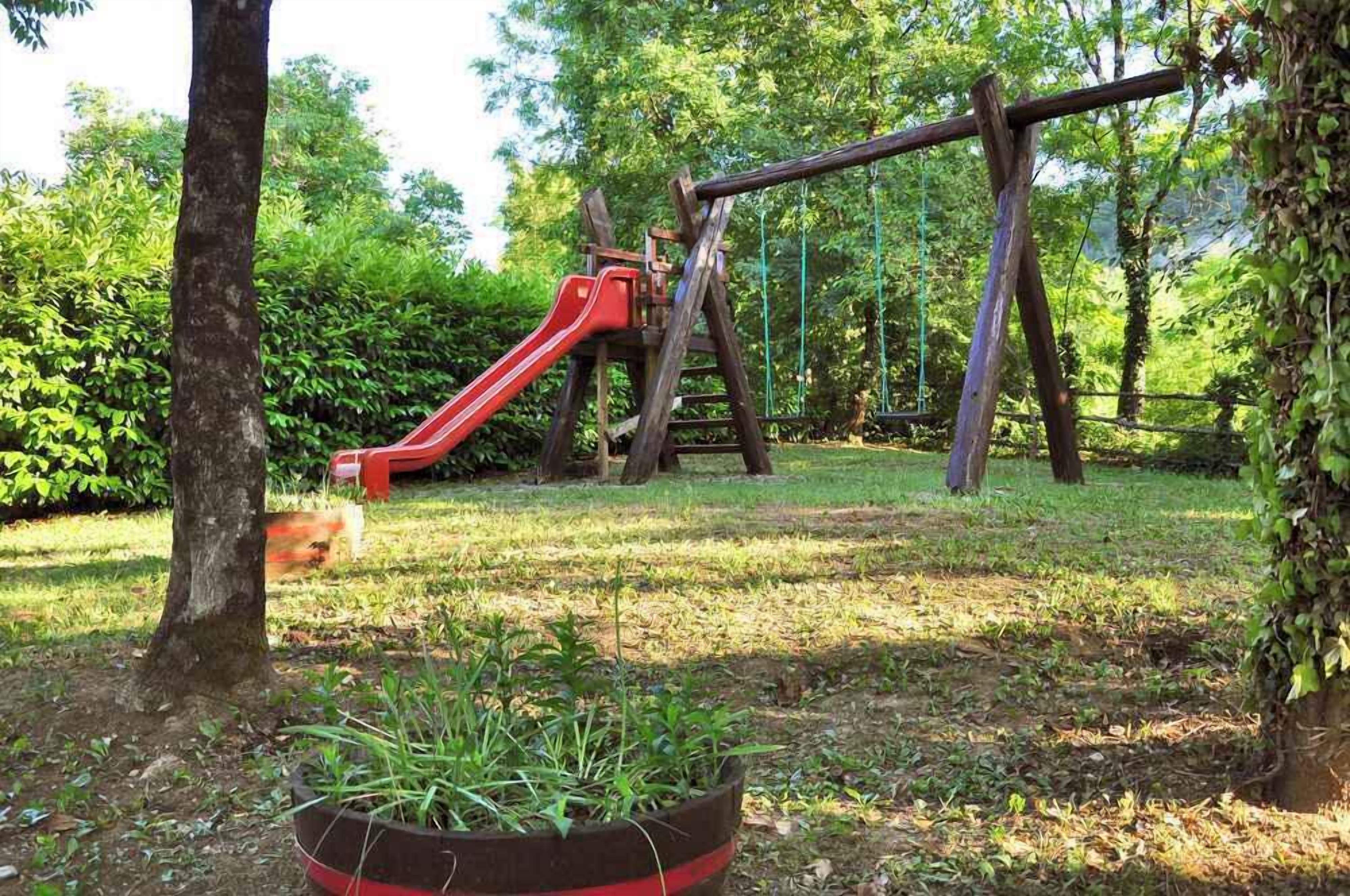 A playground with a red slide and a wooden swing set in a grassy area with trees.