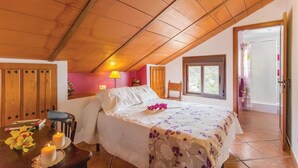 Een gezellige slaapkamer met een schuine kap, een bed met een bloemenbedekking, een nachtkastje met kaarsen, en een raam met uitzicht op groen.