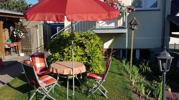 A patio with a table and chairs, a red umbrella, and a house in the background. - Apartment in Ahlbeck - Apartment (basement up to 3 pers.) (Heringsdorf)