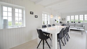 Une salle à manger spacieuse, avec une longue table blanche et des chaises noires, entourée de murs recouverts de panneaux blancs et de grandes fenêtres.
