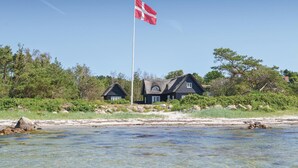 Une maison située sur la côte, avec un mât sur lequel flotte un drapeau, entourée d’arbres et donnant sur une plage.