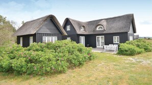 Une maison traditionnelle à toit de chaume, à l’extérieur sombre, entourée d’une végétation luxuriante et de buissons en fleurs.