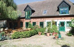 Un bâtiment traditionnel en briques, avec des encadrements de fenêtres verts, une table en bois et des bancs dans la cour, ainsi que des plantes en pot disposées tout autour.