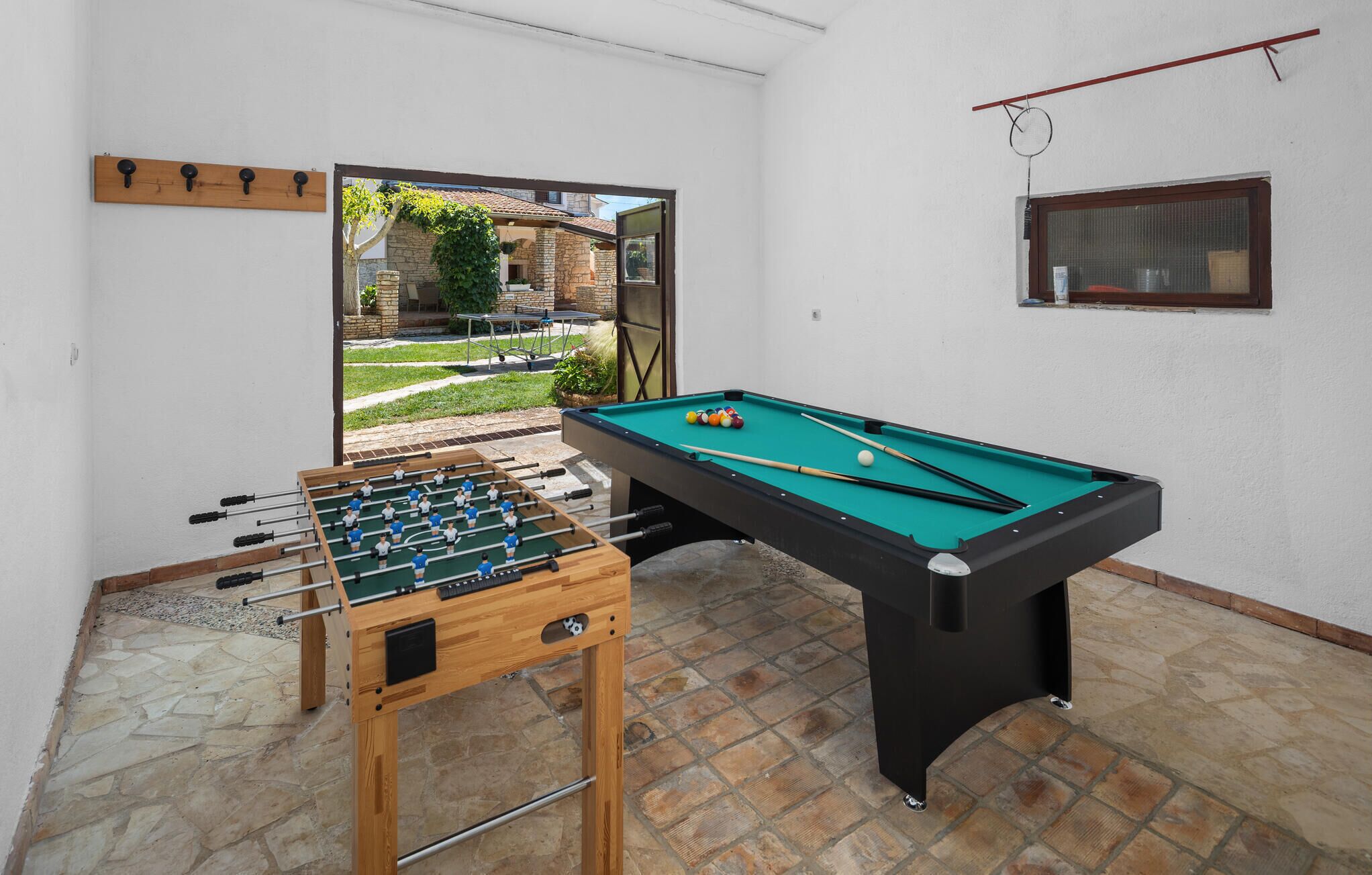 A pool table and foosball table in a room with a stone floor and a view of a garden.