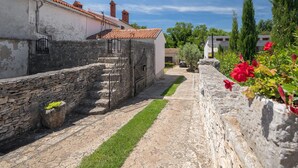 Un étroit sentier de pierre, bordé de murs en pierre, mène à un bâtiment doté d’un toit en tuiles et d’un petit escalier.