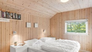 A wooden-paneled bedroom with a bed, a nightstand, a shelf with books, and a window with a view of greenery.