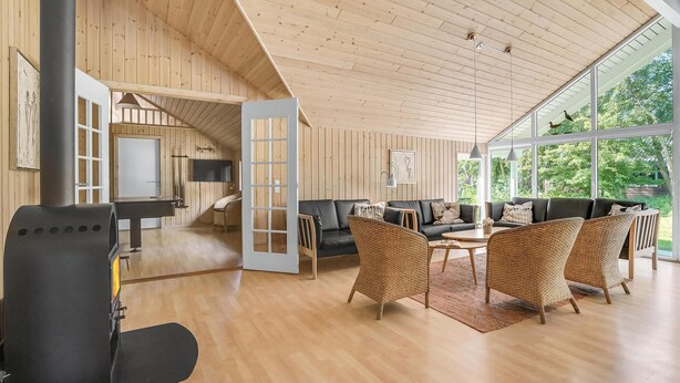 A spacious living area with a wooden ceiling, a black sofa, wicker chairs, and a large window offering a view of greenery.