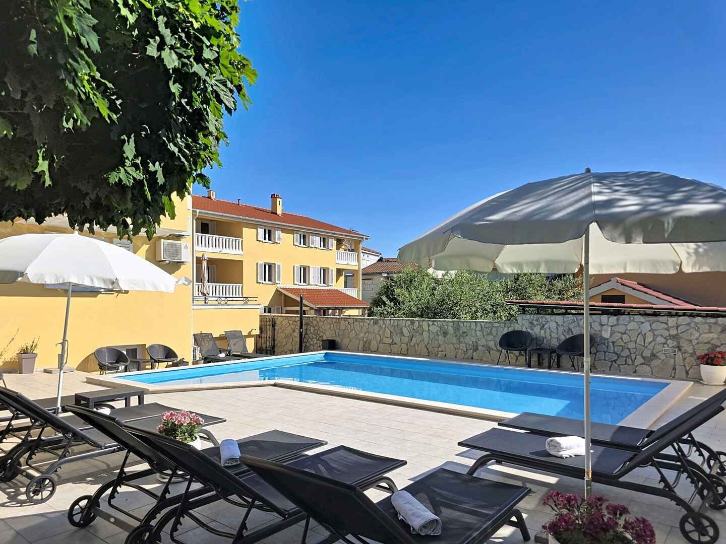 Ett poolområde med solstolar och parasoller, en stenmur och en gul byggnad med balkonger.
