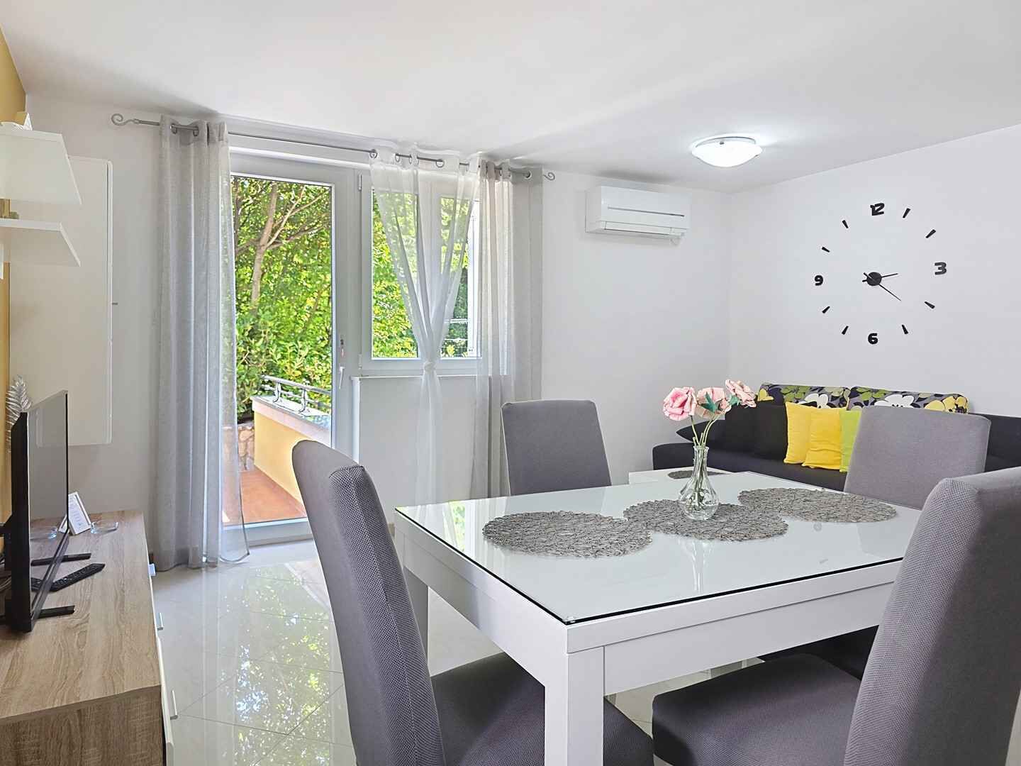 A modern living room with a glass dining table, grey chairs, a sofa with yellow cushions, a clock on the wall, and a view of greenery outside.