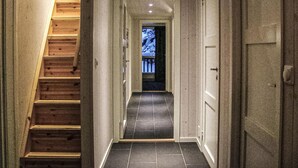 Un couloir étroit, avec un escalier en bois sur la gauche, un sol carrelé et une porte sur la droite.