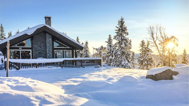 Un chalet enneigé, doté d’une cheminée en pierre, d’une terrasse en bois et offrant une vue sur des arbres couverts de neige.