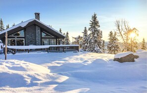 Un chalet enneigé, doté d’une cheminée en pierre, d’une terrasse en bois et offrant une vue sur des arbres couverts de neige.