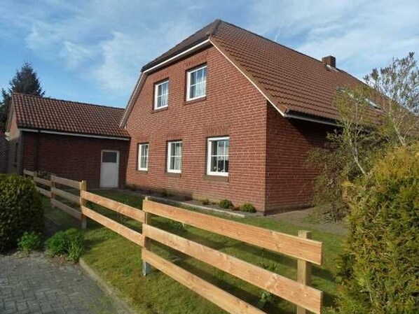 A two-story brick house with a sloped roof, a wooden fence, and a paved driveway. - Holiday apartment - Friedel (Großheide)