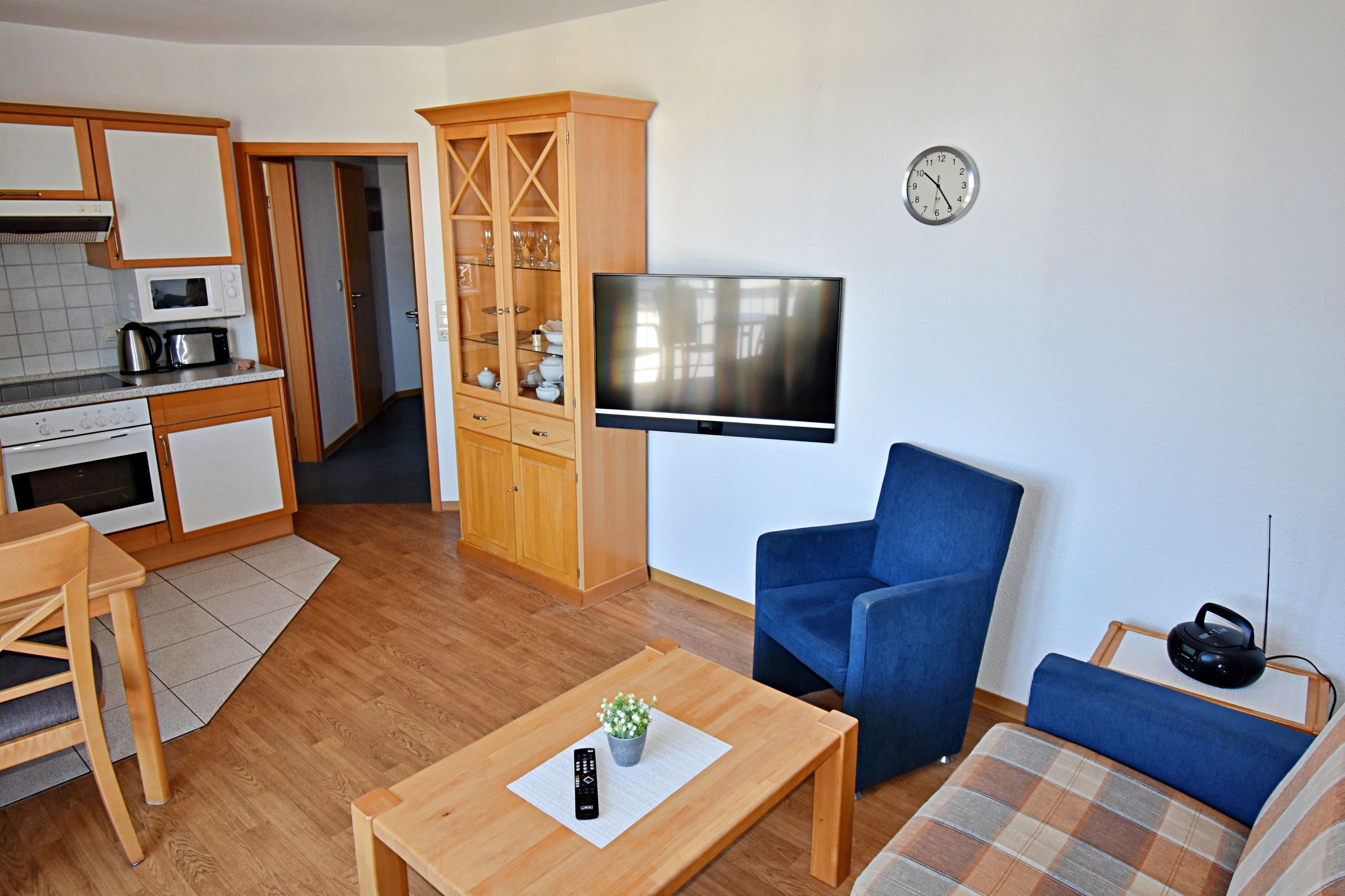 A compact living space with a kitchenette, a flat-screen TV, a blue armchair, a sofa, and a small table with a remote control.