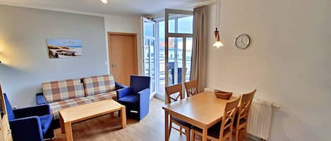 Una sala de estar con sofá, una mesa de comedor y sillas. Se ve una ventana con cortinas, una puerta y un reloj en la pared.