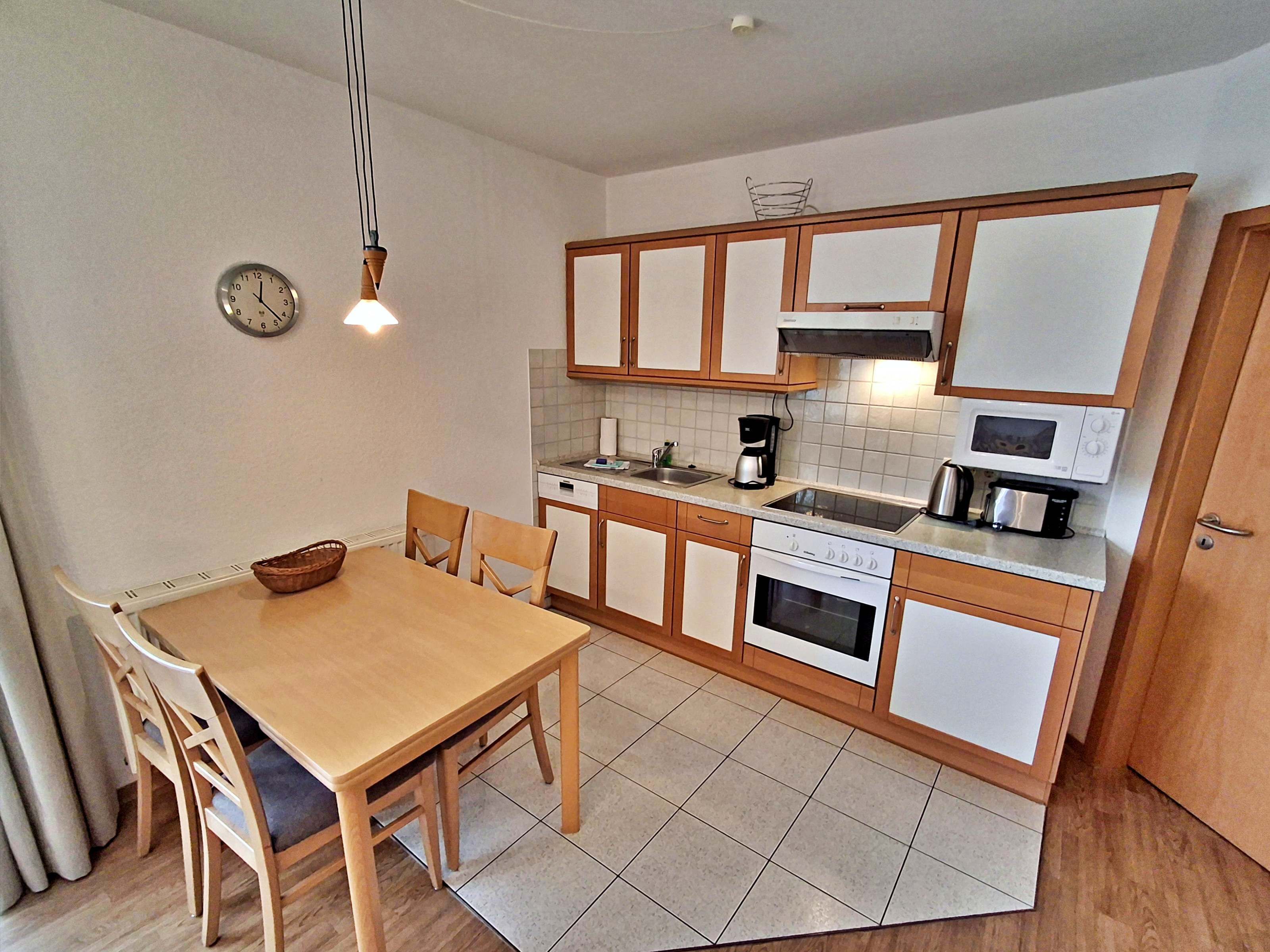 Una cocina compacta con armarios de madera, una mesa de comedor con sillas, un reloj y un microondas.