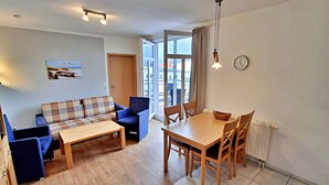 Una sala de estar con sofá, una mesa de comedor y sillas. Se ve una ventana con cortinas, una puerta y un reloj en la pared.