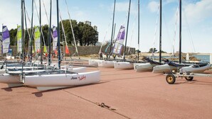 Ein Yachthafen mit mehreren Segelbooten, von denen einige Flaggen zeigen, und ein Strand im Hintergrund.