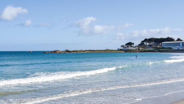Ein Strand mit klarem, blauem Wasser, Wellen und einer felsigen Landzunge im Hintergrund.