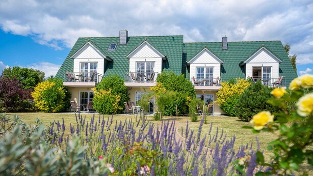 Une grande maison à plusieurs étages, dotée d’un toit végétalisé et de boiseries blanches, entourée d’un jardin bien entretenu où poussent diverses plantes et fleurs.