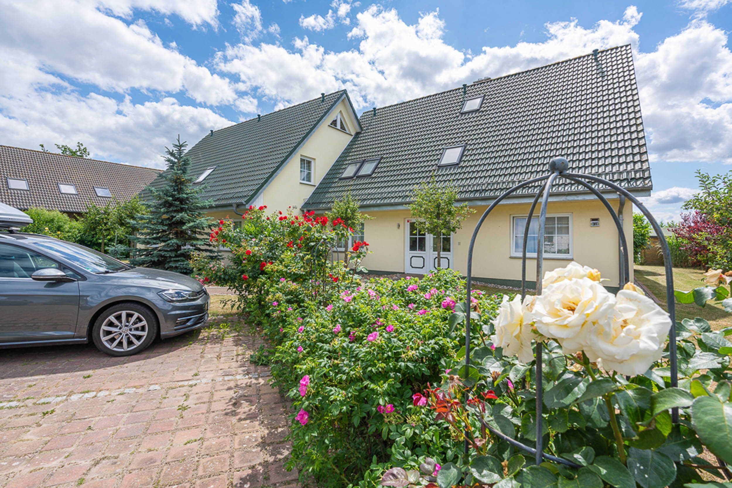 Een woonhuis met een verharde oprit, een geparkeerde auto en een tuin met bloemen.