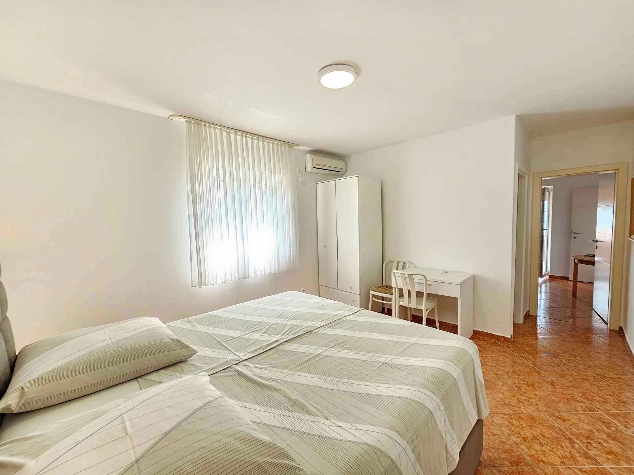 Un dormitori amb un llit, un escriptori i una cadira. Hi ha una finestra amb cortines, un aparell d'aire condicionat i una porta que dona a una altra habitació.
