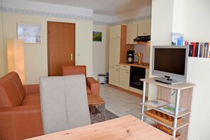 Un espace de vie compact comprenant un canapé, une petite table à manger, un téléviseur posé sur un meuble en bois, et un coin cuisine avec des placards et des appareils électroménagers.