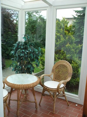 Une véranda avec une table et des chaises en osier, une plante en pot et une vue sur un jardin.