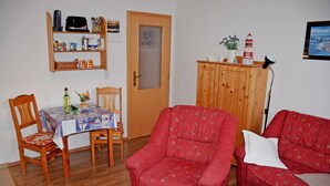 A living room with a wooden door, a dining table with chairs, a red armchair, a red sofa, and a cabinet with decorative items. - Vacation apartment Mönchgut - Holiday apartments in Gager on Rügen (Gager)