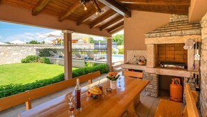 An outdoor patio with a wooden table set for dining, a stone fireplace, and a view of a swimming pool and buildings in the background. - Beautiful apartment in Hr-52215 Vodnjan (Hr-52215 Vodnjan)