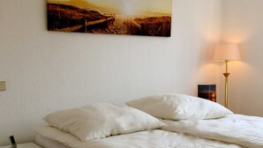 A neatly made bed with white linens, a bedside table with a lamp, and a painting of a beach scene on the wall. - Baltic Sea residence, App. 2 - Biting Resence 2-Zi.-Fewo., EC (Neustadt in Holstein)