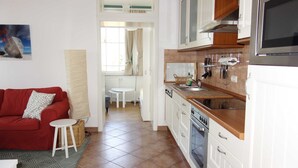 Cocina compacta con armarios blancos, encimera de madera y horno empotrado. Cuenta con un comedor con mesa y sillas blancas, además de un sofá rojo con cojín estampado.