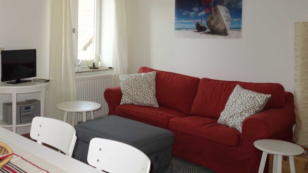 Una habitación con un sofá rojo, dos sillas blancas, una mesita, un televisor y un cuadro de barcos en la pared.
