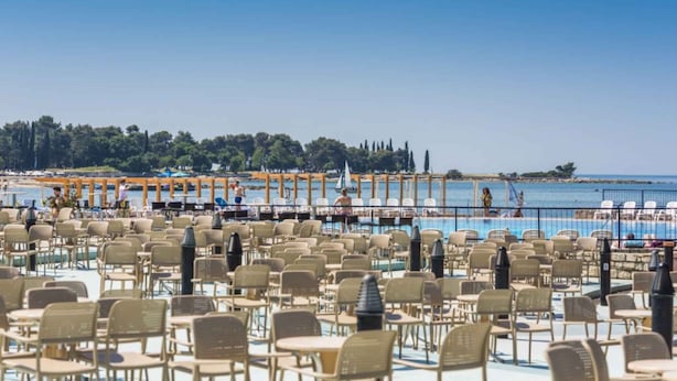 A poolside area with numerous chairs and tables, a clear view of the sea, and a distant shoreline with trees and buildings. - Holiday apartment with outdoor pool (Umag)