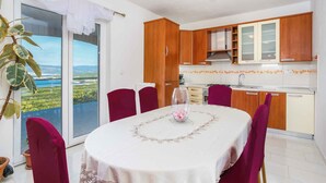 A dining area with a table set for four, a kitchen with wooden cabinets, and a view of a green landscape through a sliding door. - Holiday apartment with pool and sea view (Čižići)
