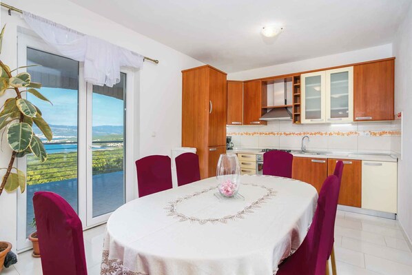 Uma sala de jantar com uma mesa posta para quatro pessoas, uma cozinha com armários de madeira, e vista para uma paisagem verde através de uma porta deslizante.