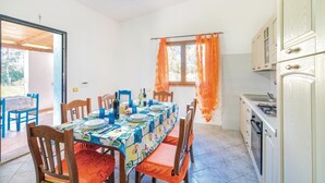 A dining area with a table set for four, wooden chairs, a window with orange curtains, and a kitchen with a stove and cabinets. - 6 bedroom beautiful home in Orosei NU (Orosei NU)
