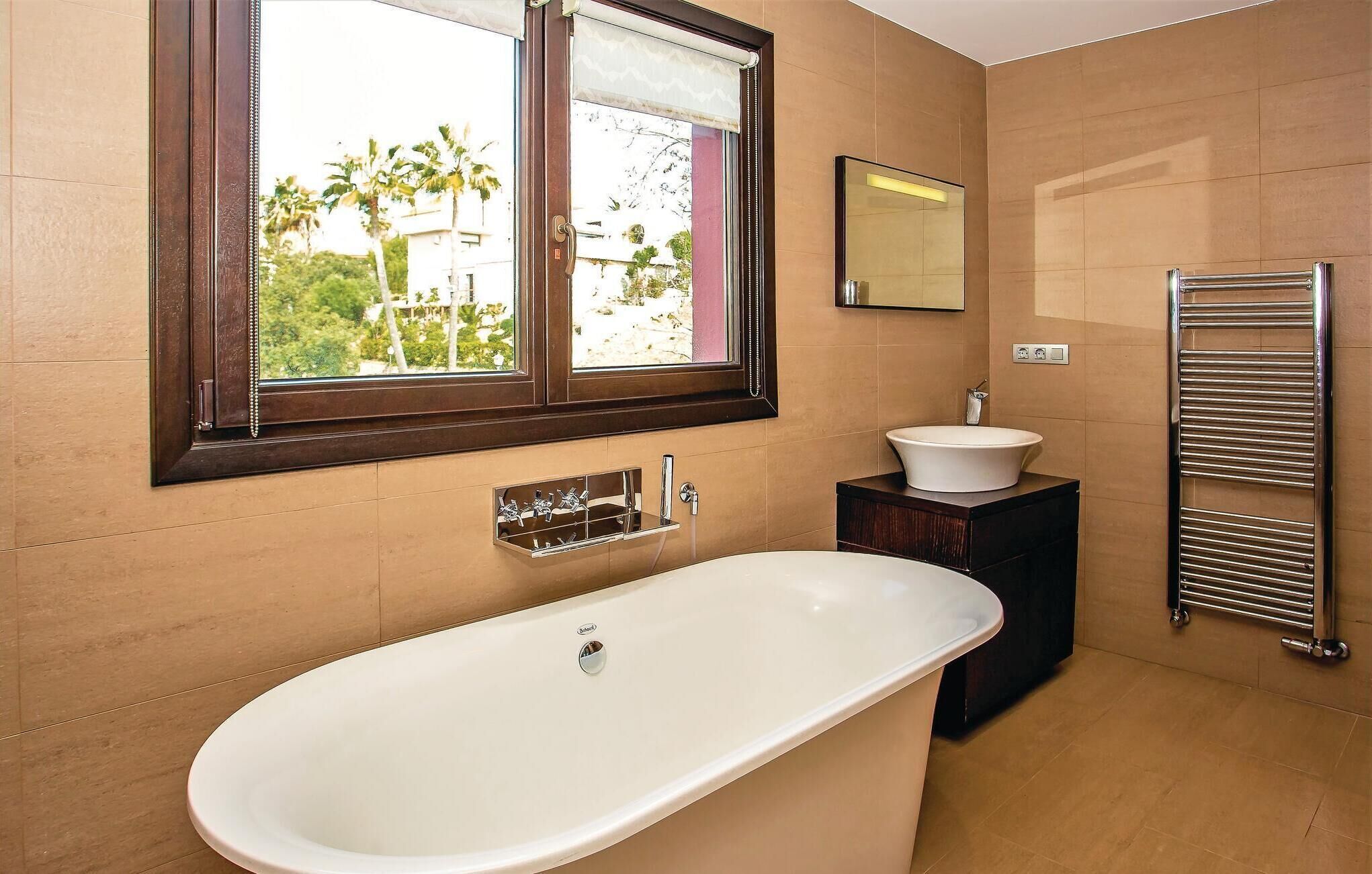 Une salle de bain avec une baignoire sur pieds, un meuble-lavabo avec un évier, un porte-serviettes et une fenêtre donnant sur des palmiers et des immeubles.