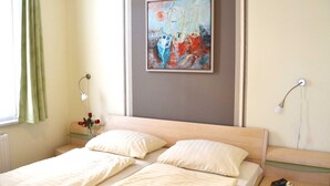 A double bed with two pillows, a bedside table with a lamp, a framed painting on the wall, and a window with curtains. - Category I, EC, 2 P. - Apartments Villa "On the sunny beach" (Heringsdorf)