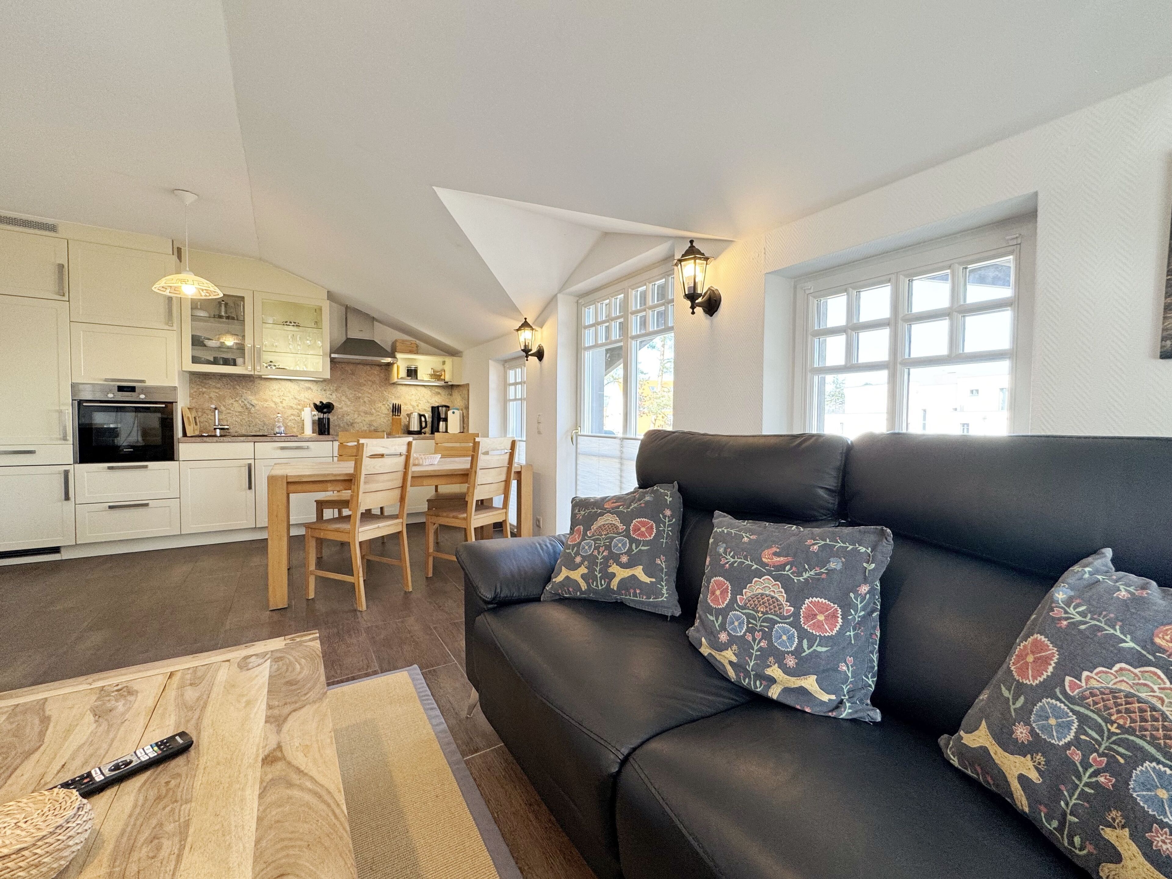 Une cuisine moderne avec des meubles blancs, un coin repas avec des chaises en bois et un canapé en cuir noir agrémenté de coussins à motifs.