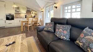 A modern kitchen with white cabinets, a dining area with wooden chairs, and a black leather sofa with patterned cushions.