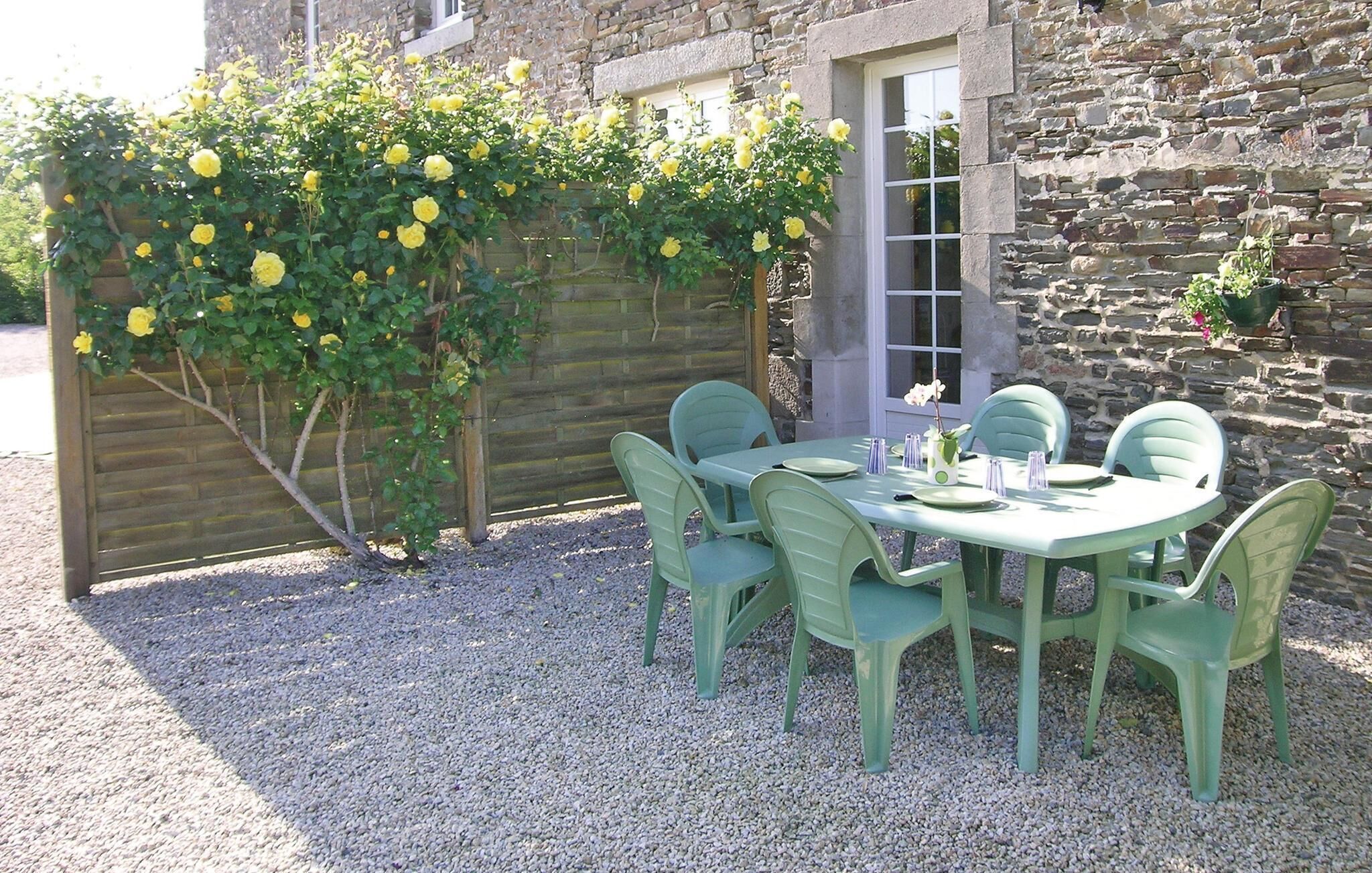 A table set outdoors with green chairs, a stone building in the background, and a yellow flowering plant.