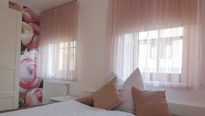 Une chambre à coucher avec un lit, des oreillers, une table de chevet, un radiateur et des rideaux légers.
