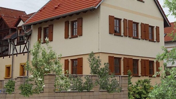 Un bâtiment à deux étages, avec un toit recouvert de tuiles rouges, de nombreuses fenêtres dotées de volets en bois et un mur de briques à l’avant.