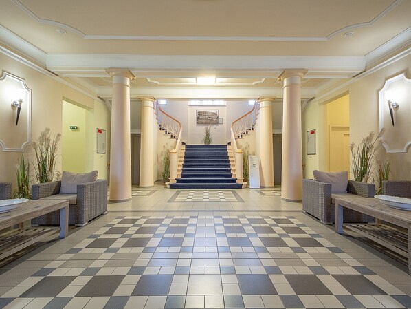 Un hall d’entrée avec un sol à carreaux, un escalier et deux espaces de détente agrémentés de plantes.