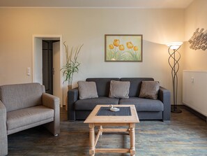 Un salon avec un canapé gris, un fauteuil gris plus petit, une table basse en bois ornée d’un motif de cygne, un lampadaire et un tableau représentant des fleurs jaunes accroché au mur.