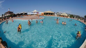 Una gran piscina al aire libre con gente nadando y jugando, rodeada de sombrillas y sillas de descanso.